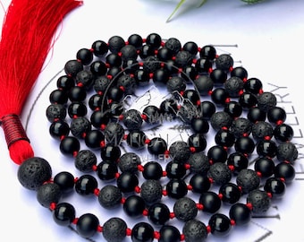 May include: A black beaded mala necklace with red string and a red tassel. The necklace features a mix of smooth, matte black beads and textured, porous lava beads. The tassel is a vibrant red.
