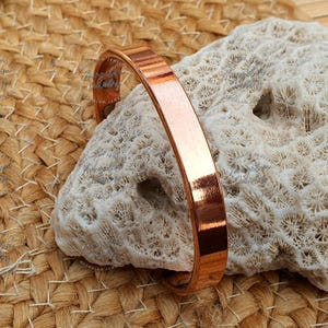 May include: A polished copper cuff bracelet. The bracelet has a flat, rectangular design and a smooth, reflective surface. The copper color is a warm, reddish-brown hue. The bracelet is displayed on a textured, off-white coral-like object.
