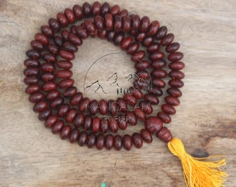 May include: A strand of dark red wooden beads, likely a prayer mala, with a yellow tassel. The beads are oval-shaped and strung together. The image includes the text "MANGALAM JEWELER".