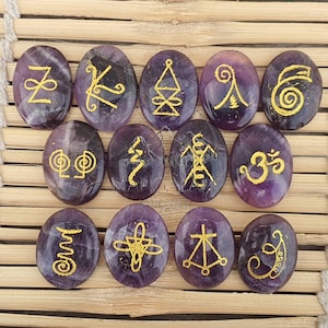 May include: Twelve oval amethyst stones, each featuring a distinct gold symbol. The stones are displayed on a light brown woven surface. The symbols are diverse, creating a mystical appearance.