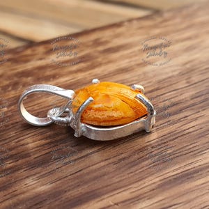 May include: A silver pendant with a large, oval, amber-colored stone. The stone is set in a silver bezel with four prongs. The pendant has a loop for a chain. The background is a wooden surface.