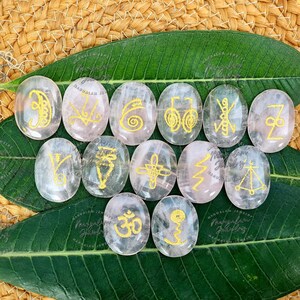 May include: A set of oval, clear quartz stones featuring gold symbols. The stones are displayed on a large green leaf and a woven, tan surface. The symbols are diverse, and the stones have a translucent quality.