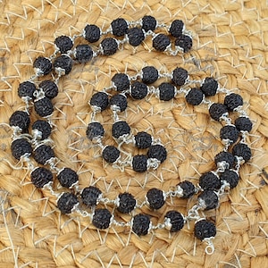 May include: A necklace composed of dark brown, textured beads strung together with silver-coloured wire. The beads are round and have a rough surface. The necklace is displayed on a woven, tan surface.