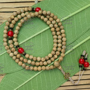 Puede incluir: Un collar mala en capas con cuentas de color marrón claro, intercaladas con detalles rojos y negros. El collar descansa sobre una hoja verde, con una borla a juego y cuentas rojas adicionales.