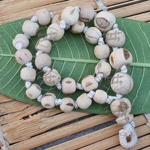 Puede incluir: Un collar hecho de cuentas de madera de color claro ensartadas en un cordón blanco. Las cuentas son redondas y cuadradas, algunas muestran la veta natural de la madera. El collar descansa sobre una hoja verde y una superficie de madera. El texto "Mangalam Jewelry" es visible.