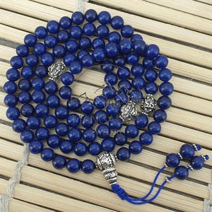 May include: A strand of deep blue gemstone beads, likely lapis lazuli, forms a necklace or prayer beads. Silver-colored decorative beads are interspersed throughout. The necklace ends with a blue tassel and is displayed on a bamboo mat.