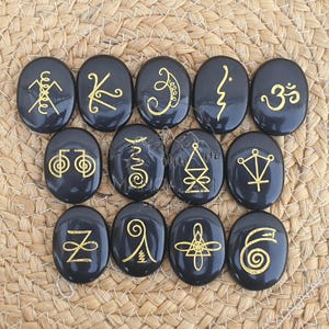 May include: Twelve black, oval stones with gold symbols. Each stone features a different gold-coloured symbol, including lines, curves, and geometric shapes. The stones are arranged in three rows of four. The stones are displayed on a woven, tan surface.