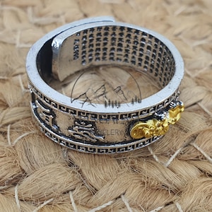 Könnte beinhalten: Silberfarbener Ring mit aufwendigen Details. Der Ring zeigt eingravierte chinesische Schriftzeichen und einen goldfarbenen Drachenakzent. Das Design umfasst eine Reihe kleiner, sich wiederholender Zeichen um das Band, vor einem strukturierten Hintergrund.
