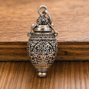 May include: An oxidized silver pendant shaped like a small, ornate urn or vessel. The surface features intricate, embossed floral and scrollwork patterns. It has a screw-top lid with a small loop at the top for hanging, and a rounded base.