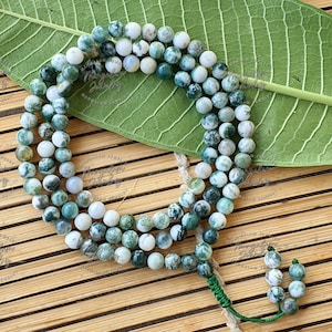 May include: A necklace of tree agate beads in shades of green and white, arranged in three rows. The beads are round and have a speckled pattern. The necklace is displayed on a green leaf and a bamboo mat.
