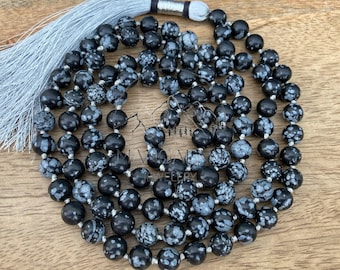 May include: A black snowflake obsidian mala bead necklace with a silver tassel. The beads are round and have a speckled pattern of white and gray. The necklace is strung on a silver cord.