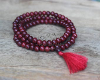 May include: A maroon mala bead necklace with a red tassel. The beads are made of wood and are strung together. The necklace is displayed on a wooden surface. The brand name "MANGALAM JEWELRY" is visible.