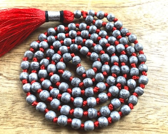May include: A silver grey mala with red string and a red tassel. The mala is made of round beads and is approximately 108 beads long.