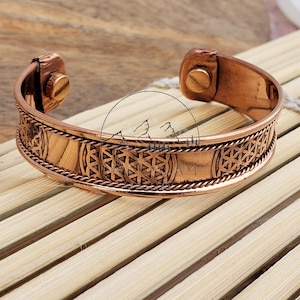 Puede incluir: Una pulsera de cobre con un intrincado diseño geométrico. La pulsera presenta una serie de patrones repetidos y un borde texturizado en forma de cuerda. El metal tiene un tono marrón rojizo cálido, y el cierre es visible en ambos extremos.