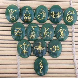 May include: Twelve oval green stones, each featuring a different gold symbol. The symbols include spirals, geometric shapes, and lines. The stones are displayed on a light brown woven surface. Each stone is approximately 3 cm in length.