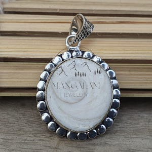 Puede incluir: Colgante de plata con un centro redondo y blanco con un diseño de montaña y árboles. El colgante tiene un borde texturizado y una fianza decorativa. El texto "MANGALAM JEWELRY" está impreso en el colgante.