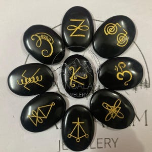 May include: Nine black, oval stones with gold symbols. The stones are arranged in a circle. The symbols are varied, featuring lines, curves, and geometric shapes. The stones are likely used for divination or spiritual practices, similar to runes or other tools.