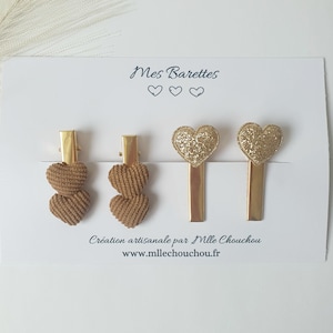 以下が含まれることがあります： 金色のハート型のヘアクリップ5個セット。2つのクリップは茶色のコーデュロイのハートのデザイン、3つのクリップは金色のグリッターのハートのデザインです。クリップは、「Mes Barrettes」と「Création artisanale par Mlle Chouchou www.mllechouchou.fr」というテキストが書かれた白いカードに表示されています。