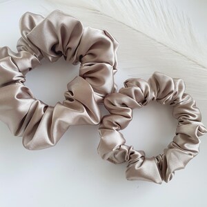 Puede incluir: Dos scrunchies de sat&eacute;n en color marr&oacute;n claro. El scrunchie m&aacute;s grande est&aacute; en primer plano y el m&aacute;s peque&ntilde;o est&aacute; detr&aacute;s.