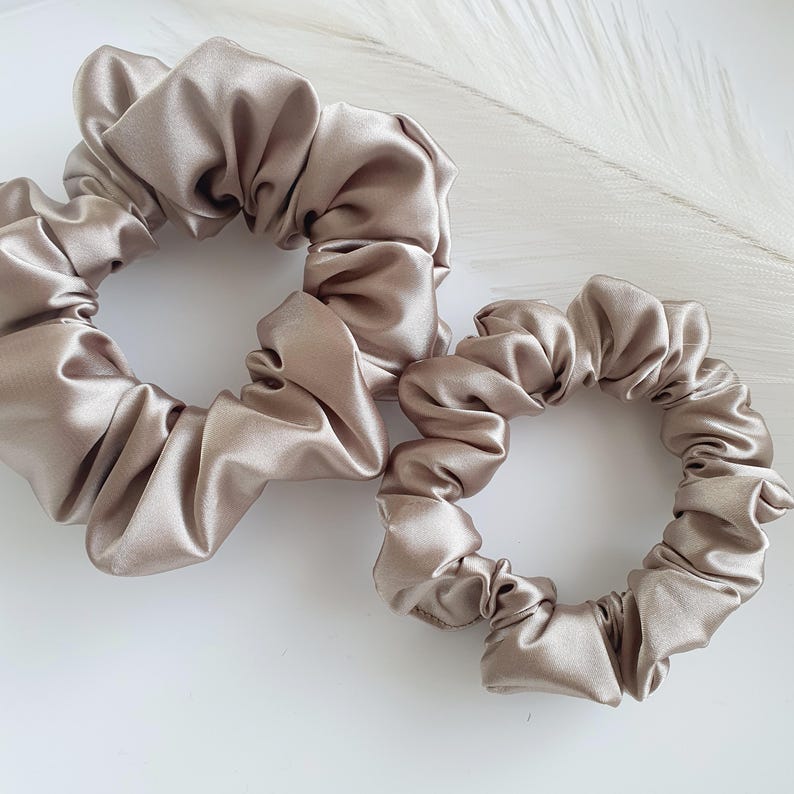 Puede incluir: Dos scrunchies de sat&eacute;n en color marr&oacute;n claro. El scrunchie m&aacute;s grande est&aacute; en primer plano y el m&aacute;s peque&ntilde;o est&aacute; detr&aacute;s.