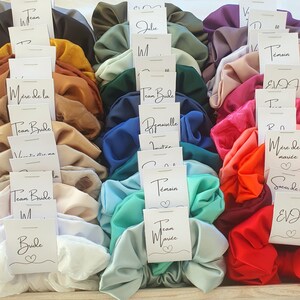 Puede incluir: Una colecci&oacute;n de scrunchies de sat&eacute;n de colores vivos con tarjetas blancas unidas a cada uno. Las tarjetas tienen diferentes textos, incluyendo "Team Bride", "Bride", "Dama de honor", "Damas de honor", "Madre de la novia" y "Hermana de la novia".