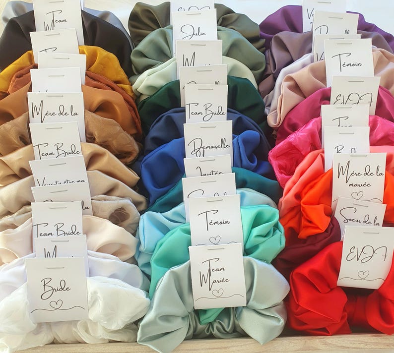 Puede incluir: Una colecci&oacute;n de scrunchies de sat&eacute;n de colores vivos con tarjetas blancas unidas a cada uno. Las tarjetas tienen diferentes textos, incluyendo "Team Bride", "Bride", "Dama de honor", "Damas de honor", "Madre de la novia" y "Hermana de la novia".