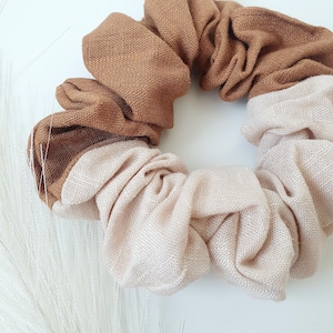 Peut inclure: Gros plan sur un chouchou bicolore. L'accessoire pour cheveux est fait d'un tissu doux, avec une couleur beige clair d'un côté et un marron moyen de l'autre. Le tissu est froncé et plié, créant un aspect texturé.