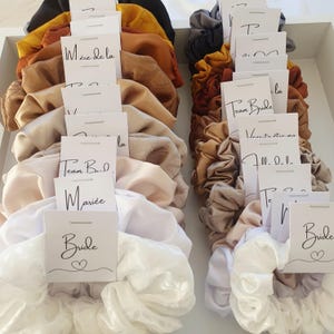 Puede incluir: Surtido de scrunchies de sat&eacute;n de colores vivos con tarjetas blancas adjuntas. Las tarjetas tienen texto escrito a mano, incluyendo "Bride", "Team Bride", "M&egrave;re de la Mari&eacute;e" y "Voulez-vous m'&eacute;pouser?"