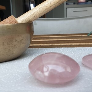 May include: Two pink rose quartz heart-shaped stones on a white surface. A brass singing bowl with a wooden striker is in the background.