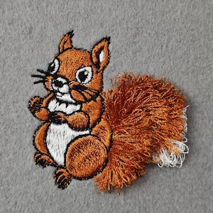 May include: Embroidered squirrel patch in brown, white, and black, with a fluffy tail. The patch is set against a grey background. Ideal for adding to clothing or accessories. The patch is approximately 7 cm in height.