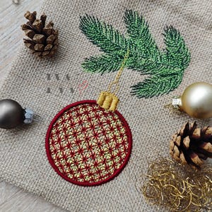 May include: Embroidered Christmas ornament with a red and gold pattern, surrounded by a green embroidered pine branch, pine cones, and gold and silver baubles on a burlap background. The ornament has a gold cap and string.