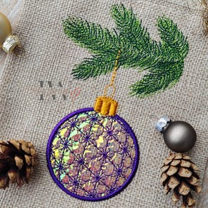 May include: Embroidered Christmas bauble with a purple and iridescent design, a gold cap, and a green branch. The bauble is on a burlap background, with a pine cone and other baubles.