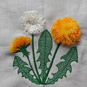 Puede incluir: Diseño bordado de tres dientes de león con hojas verdes sobre una tela de lino blanca. Los dientes de león son blancos, amarillos y naranjas.
