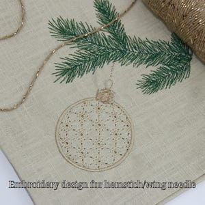 Puede incluir: Primer plano de una tela beige con un diseño bordado de adorno navideño. El adorno es beige con un patrón detallado, colgando de una rama de pino bordada verde. El texto "Embroidery design for hemstitch/wing needle" está en la parte inferior.