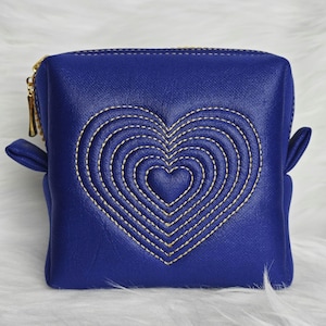 Peut inclure: Un sac cosmétique carré bleu cobalt avec un motif de cœur cousu. Le cœur est souligné de fil doré, et le sac a une fermeture éclair dorée et de petits rabats latéraux.