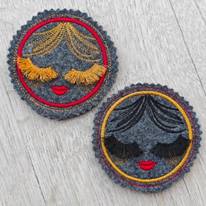 May include: Two round felt coasters with embroidered faces. One coaster has yellow eyelashes and red lips, the other has black eyelashes and red lips.