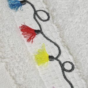 May include: Close-up of a white towel with an embroidered design. The design features a black stem with colorful tassels in blue, red, yellow, and purple. The towel has a textured surface.