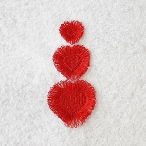 Puede incluir: Tres corazones de fieltro rojo de diferentes tamaños, apilados uno encima del otro, sobre un fondo blanco difuso.