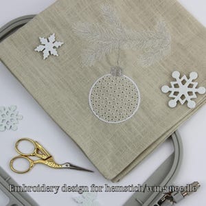 Könnte beinhalten: Beiger Leinenstoff mit gesticktem Weihnachtsbaumschmuck-Design, einem Tannenzweig und zwei weißen Schneeflocken-Dekorationen. Eine goldene Schere und ein Stickrahmen sind ebenfalls sichtbar. Der Text "Embroidery design for hemstitch/wing needle" befindet sich unten.