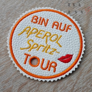 May include: A circular white patch with orange stitching and a scalloped edge. The patch has the text "BIN AUF APEROL Spritz TOUR" in orange and yellow, and a red lip graphic.