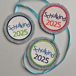 May include: Three circular felt ornaments with the text "Schulkind 2025" embroidered in blue, red, and green. Each ornament has a gray scalloped edge and a light blue ribbon for hanging.