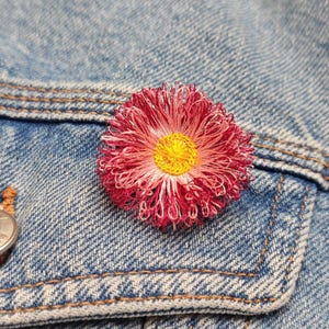 以下が含まれることがあります： ピンクと白の刺繍が施された花のブローチで、中心は黄色です。ブローチは青いデニムジャケットに留められています。