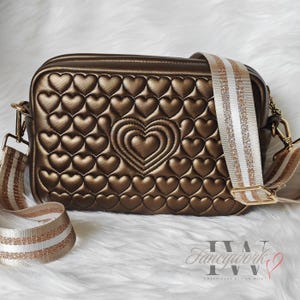 Peut inclure: Un sac bandoulière bronze avec un motif matelassé en forme de cœur. Le sac présente un grand cœur au centre et une large bandoulière rayée avec des accents dorés et blancs. La quincaillerie dorée et le texte "Fancywork IVV" sont visibles.
