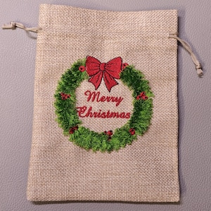 Könnte beinhalten: Eine Geschenktüte aus Jute mit Kordelzug. Die Tasche hat ein gesticktes Design mit einem grünen Weihnachtskranz mit einer roten Schleife und dem Text "Frohe Weihnachten".