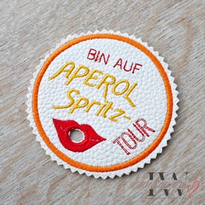 May include: Circular white coaster with an orange border and scalloped edge. The text "BIN AUF APEROL Spritz-TOUR" is embroidered in yellow and red. A red lip graphic is also featured.