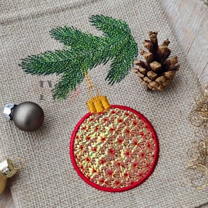 May include: Embroidered Christmas ornament with a red border and gold and red patterned center. A green embroidered pine branch and a brown pine cone are also visible. A small, dark gray ornament is on the left.