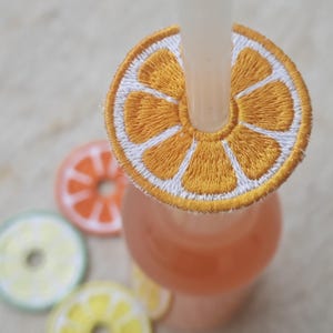 Könnte beinhalten: Ein bestickter Strohhalm-Aufsatz in Form einer Orangenscheibe sitzt auf einer Glasflasche mit rosa Flüssigkeit. Weitere bestickte Untersetzer in Form von Fruchtscheiben in Orange, Grün und Gelb sind sichtbar. Der Strohhalm ist weiß.