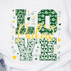 May include: White t-shirt with a green and gold "LOVE" graphic. The letters are made up of a pattern of hearts, kisses, and ribbons. The word "Packers" is written in gold letters in the center of the graphic.