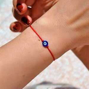 May include: A red string bracelet with a blue evil eye charm. The bracelet is worn on a person's wrist.