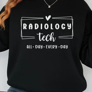May include: Black sweatshirt with the text "RADIOLOGY tech ALL DAY EVERY DAY" in white. The text is inside a white rectangle with a heart above the word "tech". The sweatshirt has long sleeves and a crew neck.
